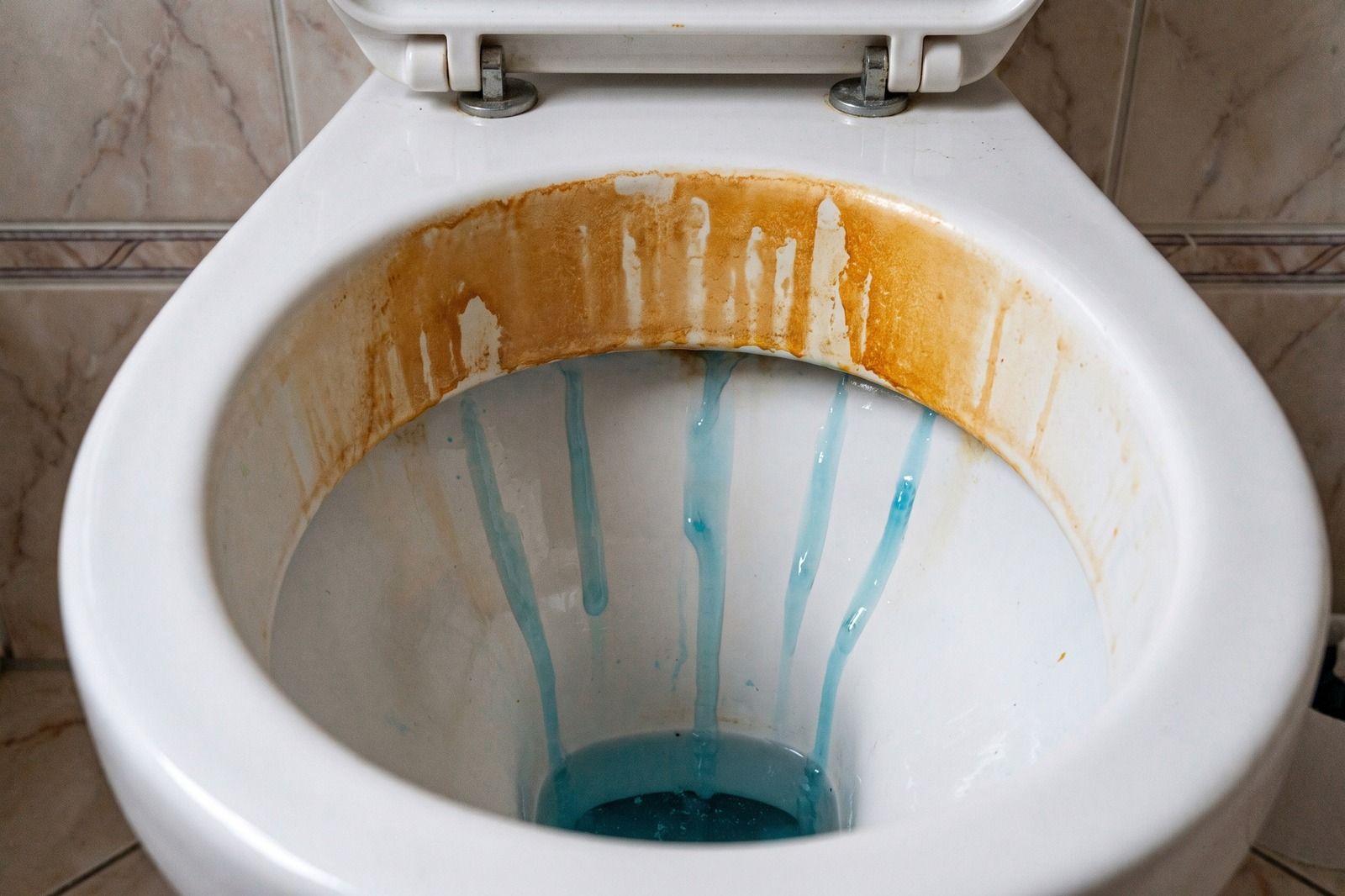 Blick in eine Toilettenschüssel: blaues Gel läuft in Bahnen an der Innenwand nach unten, oben unter der Kante bleibt ein breiter orange-brauner Belag zurück.